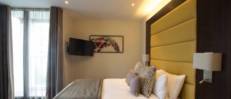 Habitación de hotel con cama, televisor montado, lámpara de pared y ventana con cortinas.