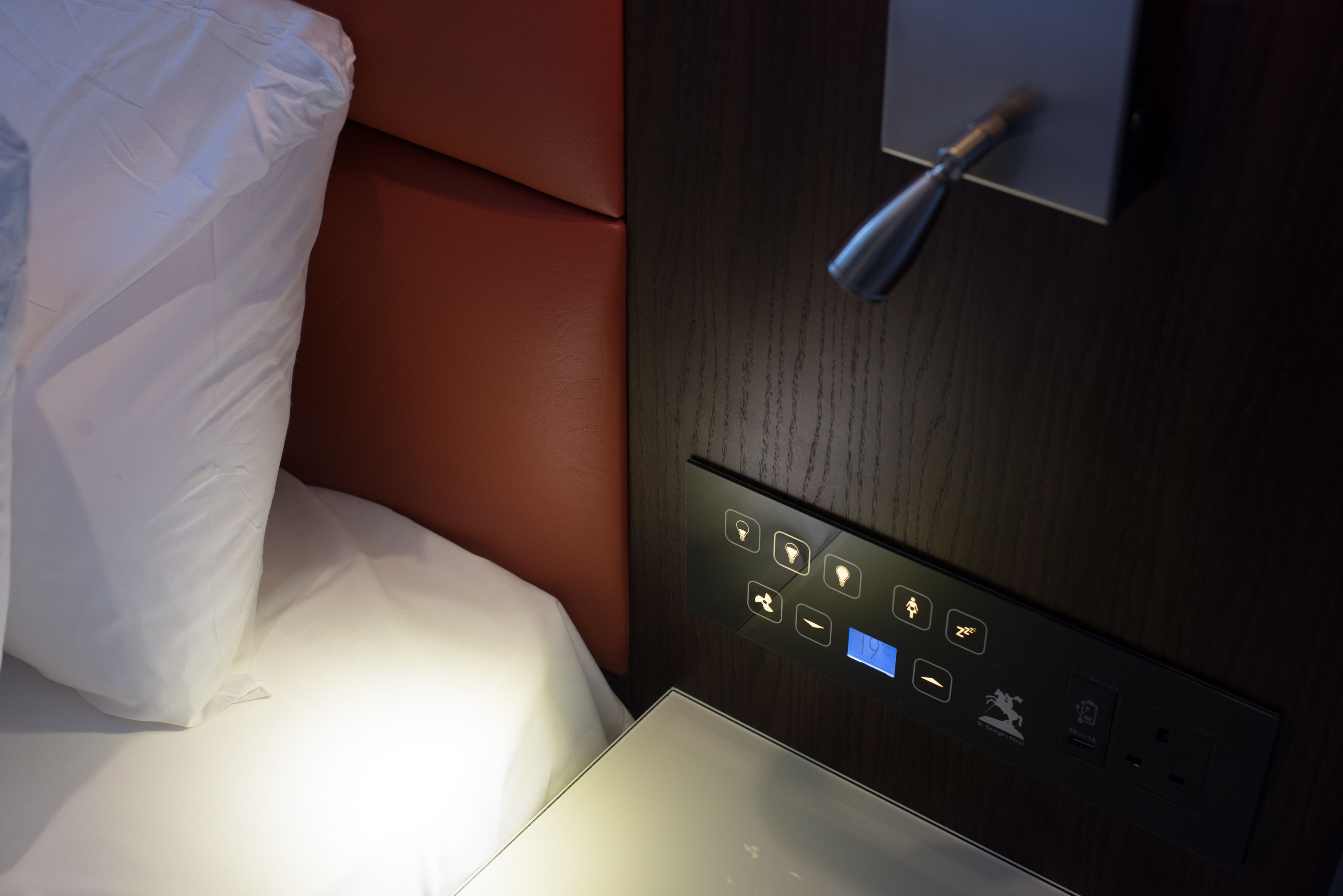A hotel room with a bed, a control panel with illuminated buttons, and a wall-mounted light fixture.