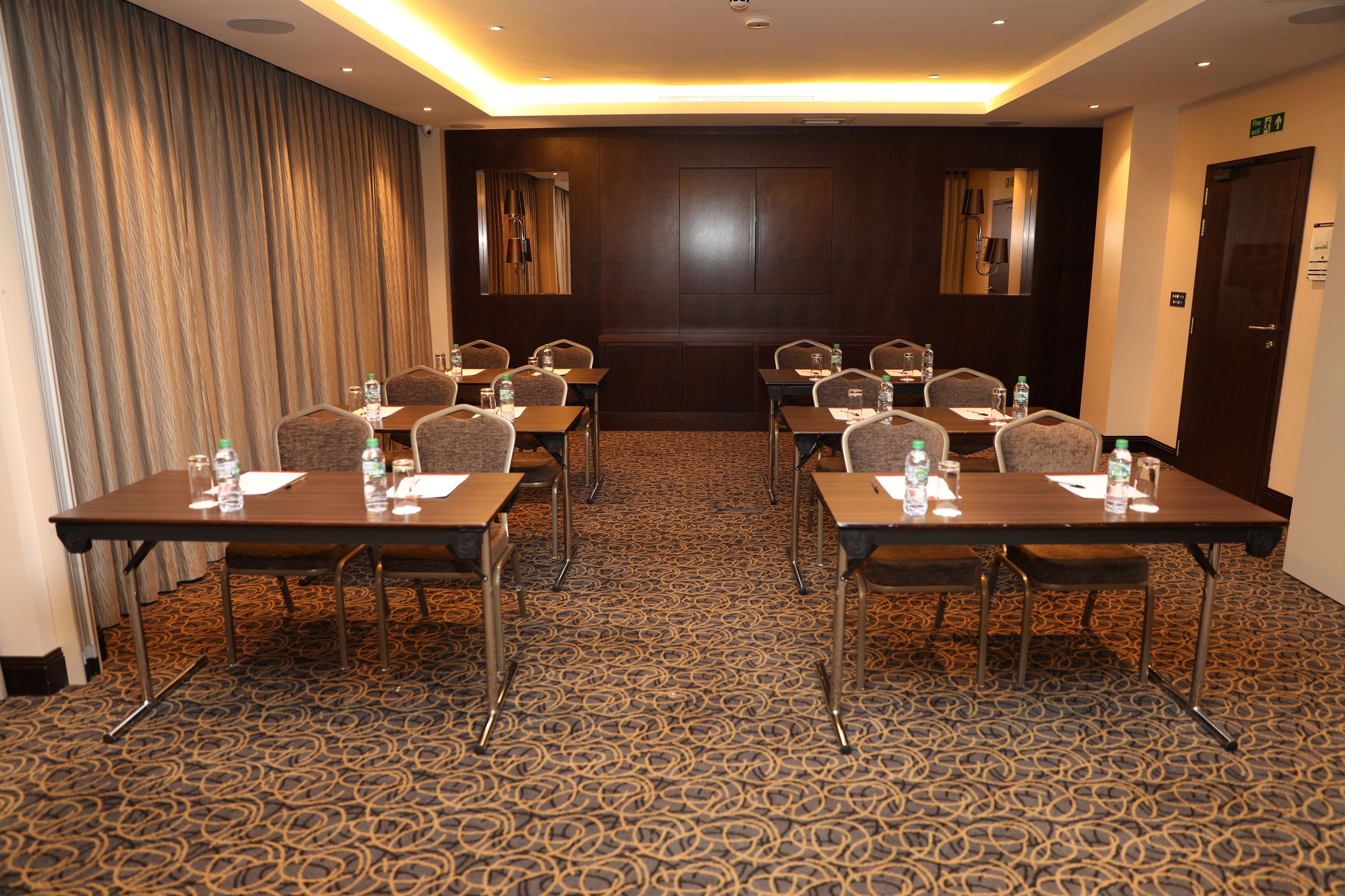 A conference room with a patterned carpet, wooden panel walls, and a door on the right. Tables are equipped with chairs, water bottles, and papers.