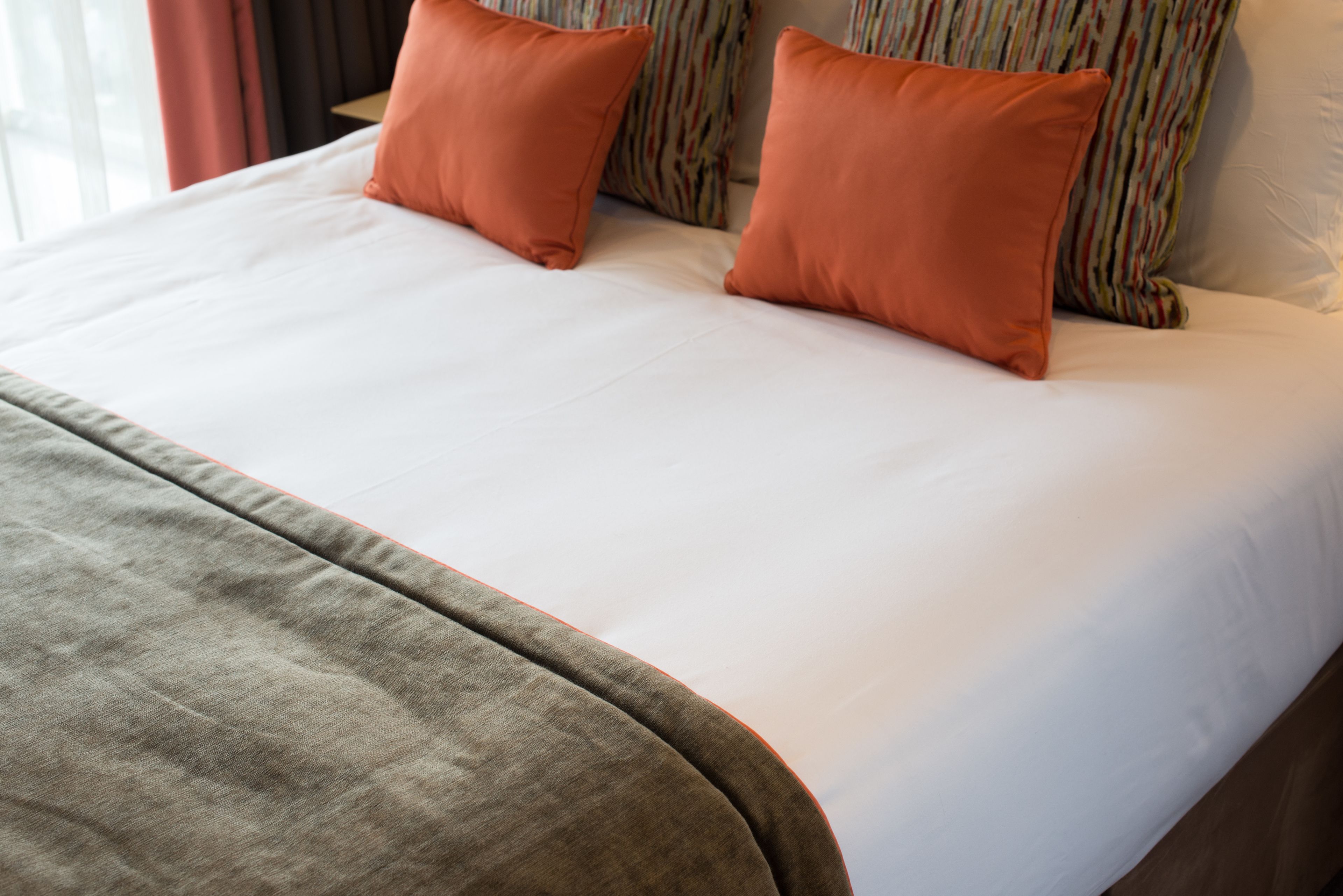Un lit bien fait, avec des draps blancs et deux oreillers orange, une couverture marron et, en arrière-plan, des oreillers à rayures.