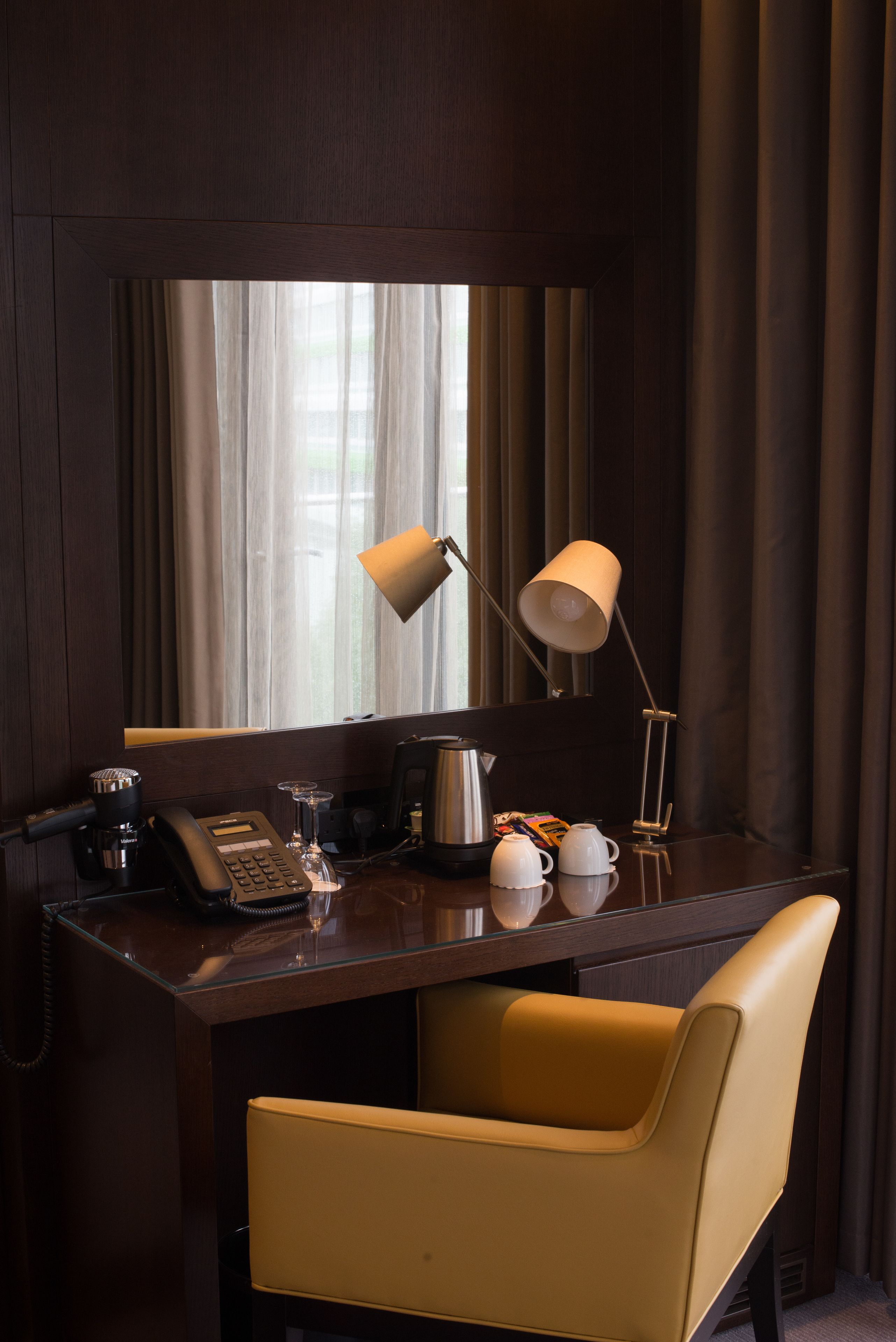 A hotel room desk with a telephone, a lamp, a coffee pot, and a chair.