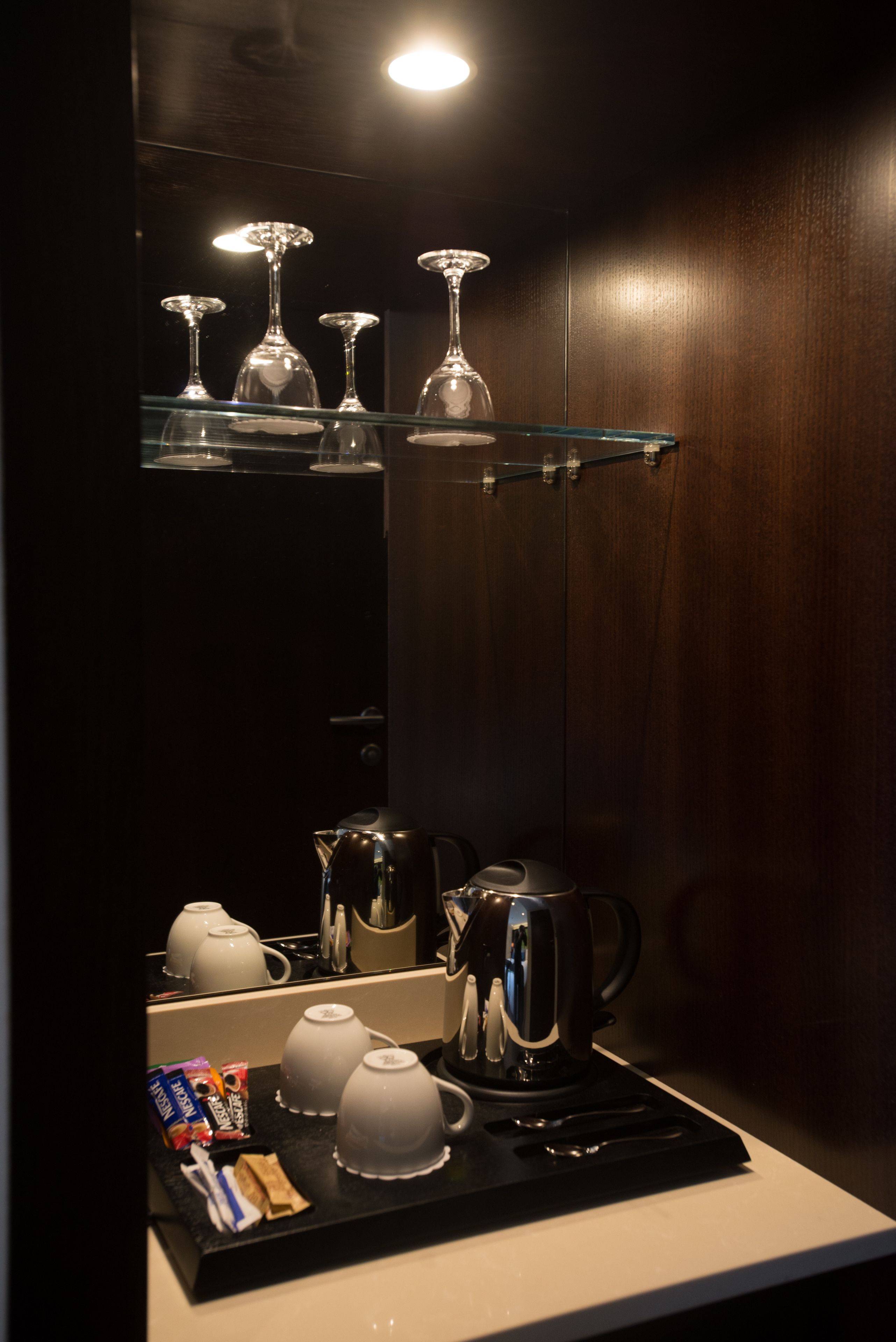 A compact kitchen with a coffee maker, teapots, and a glass shelf holding wine glasses.