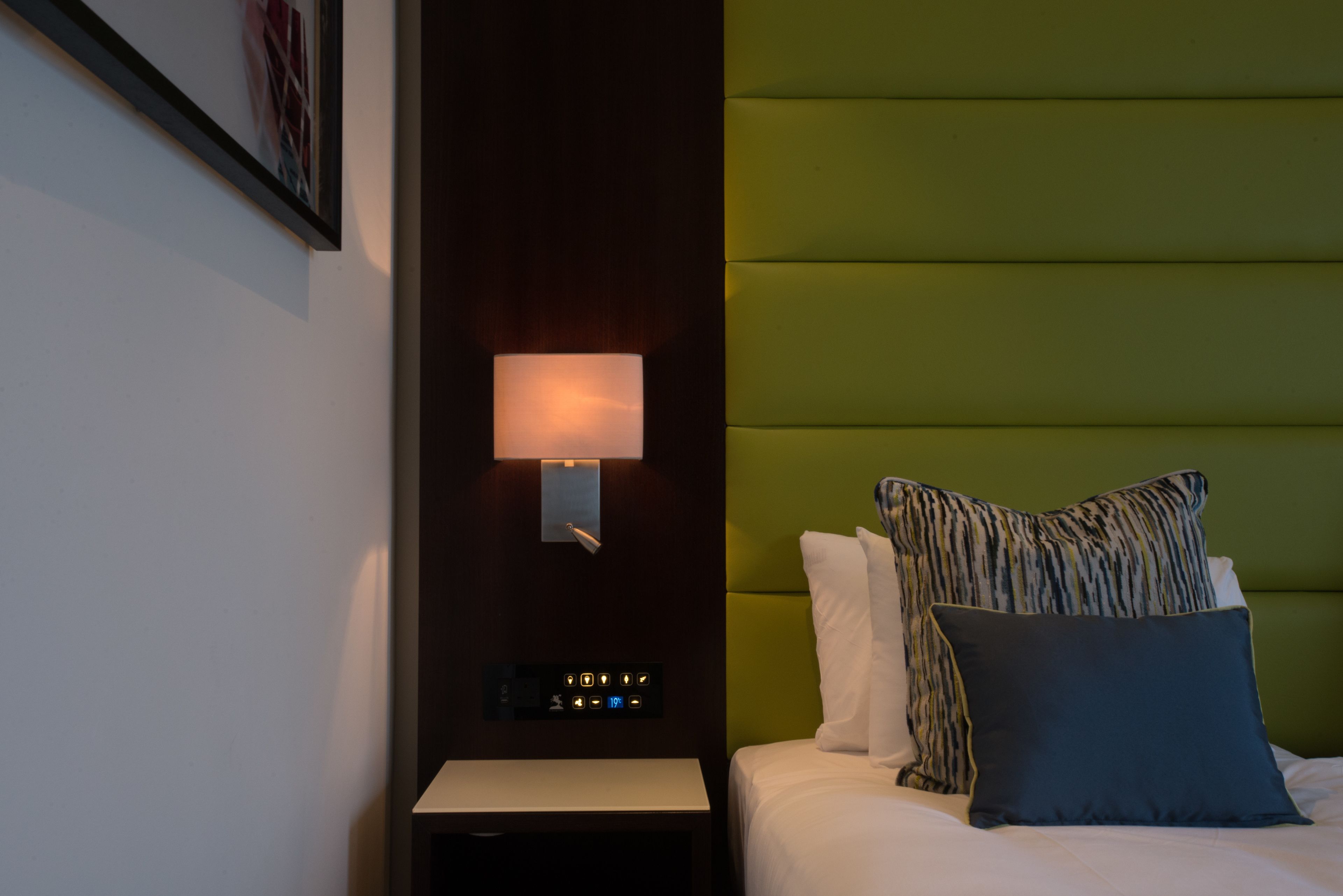 A hotel room with a bed, a nightstand, a wall-mounted lamp, and a green headboard.