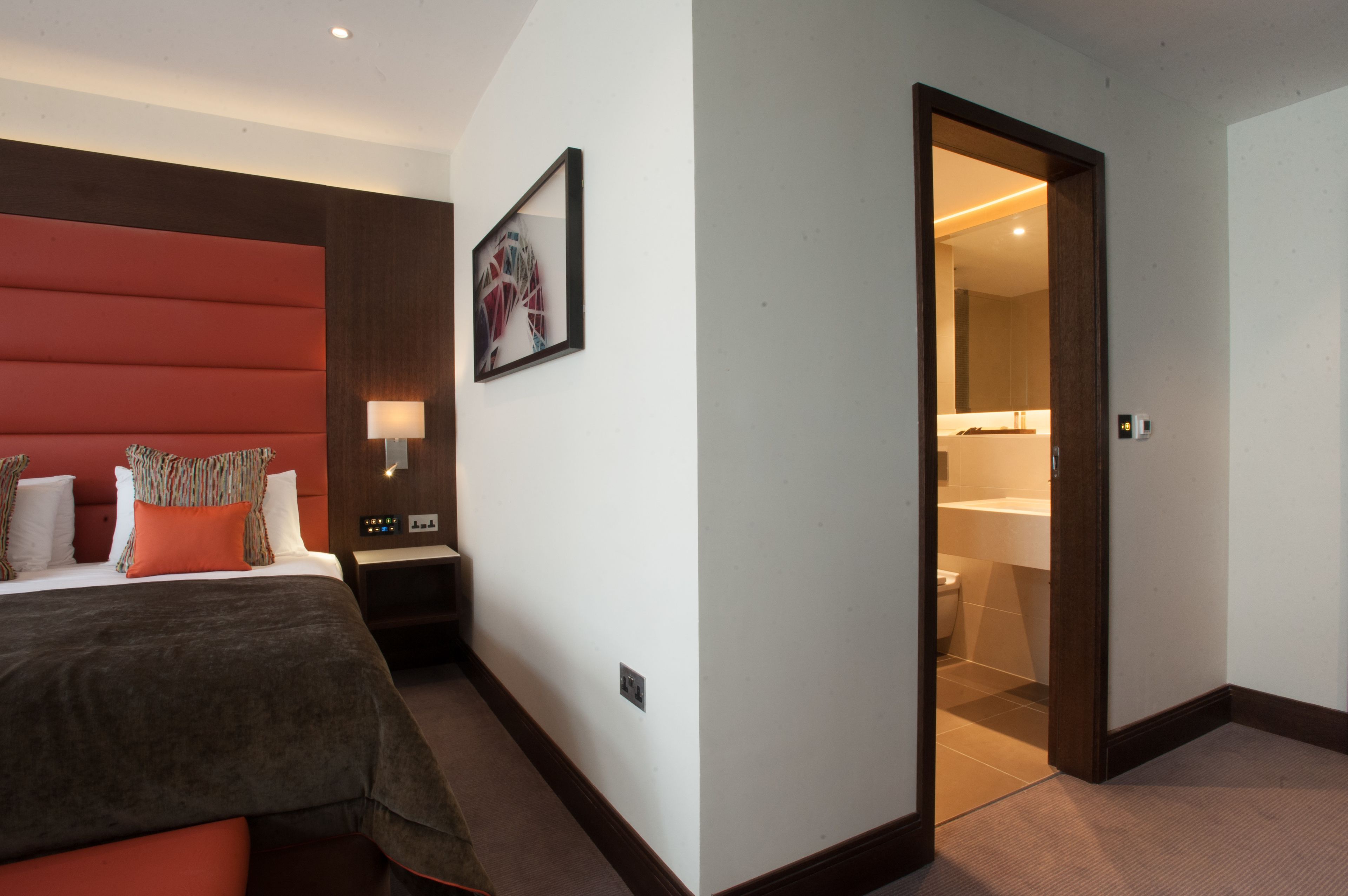 A hotel room with a large bed, a red accent wall, a painting, and a bathroom visible through an open door.