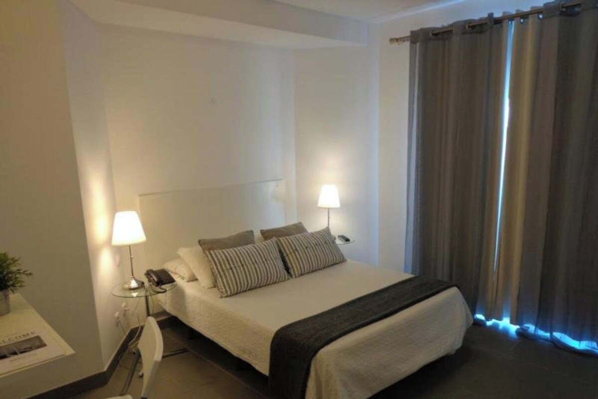 A hotel room with a bed, bedside lamps, a desk with a lamp, and a window with curtains.