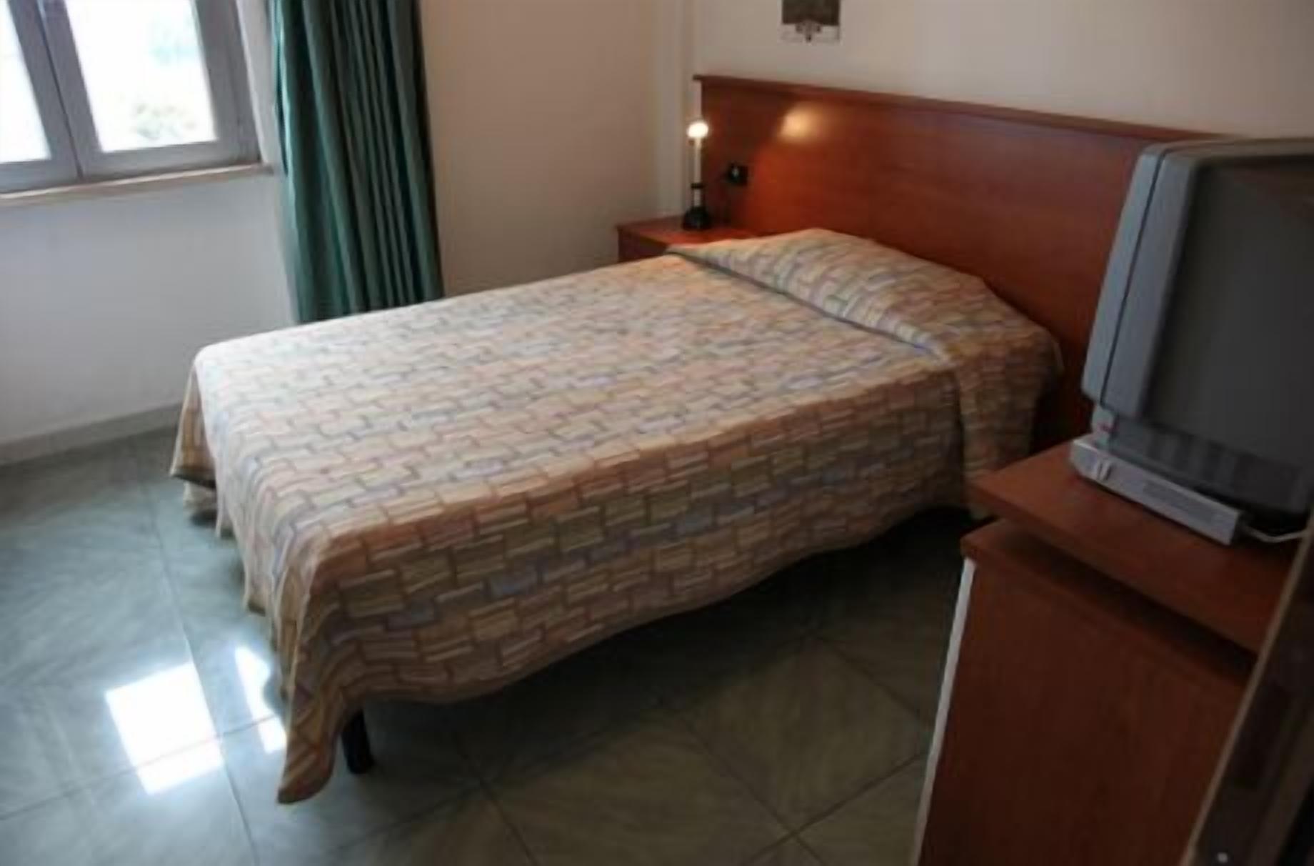 Une chambre d’hôtel avec un lit, une table de chevet avec une lampe, un téléviseur posé sur un meuble en bois et une fenêtre avec des rideaux.