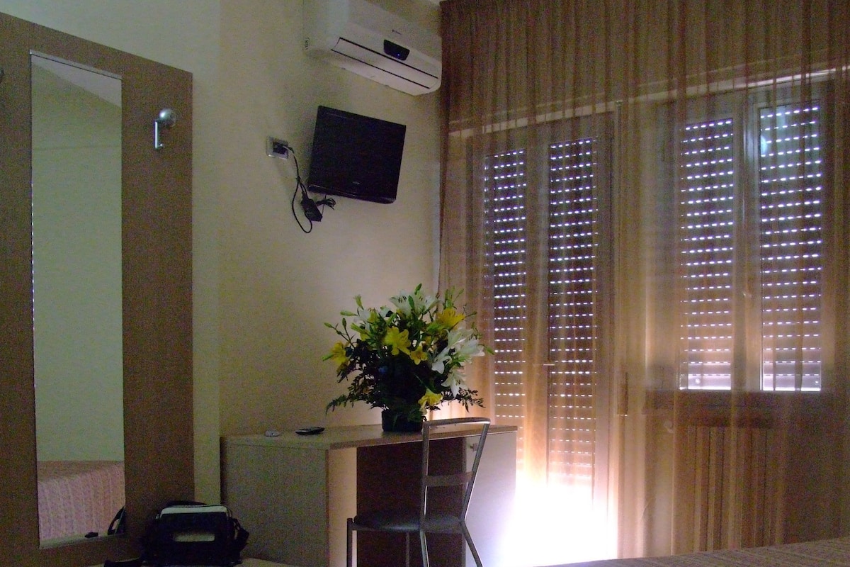 A hotel room with a bed, desk, chair, air conditioning unit, television, and a window with blinds.