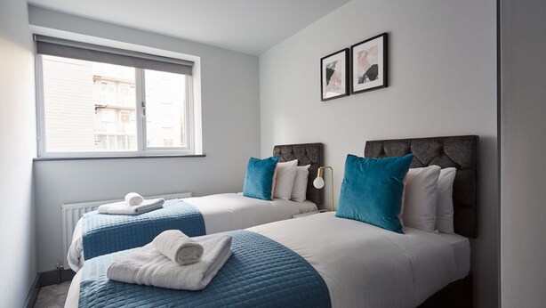 Una camera d'albergo con due letti, ognuno con una coperta trapuntata blu e lenzuola bianche. Si vede una finestra con vista su edifici, e due quadri appesi al muro.