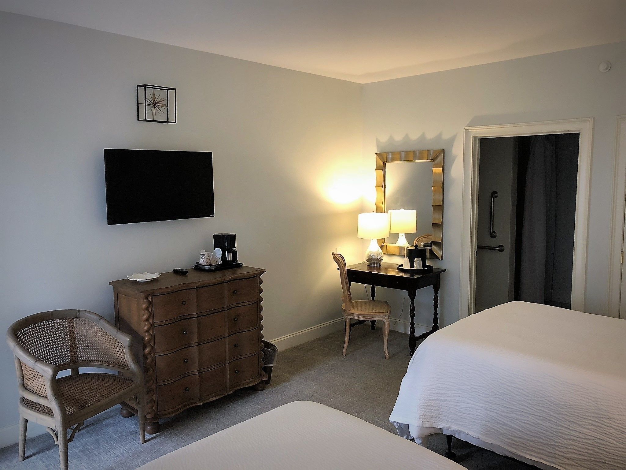 Una habitación de hotel con cama, tocador, una silla, un escritorio con lámpara, televisión y espejo.