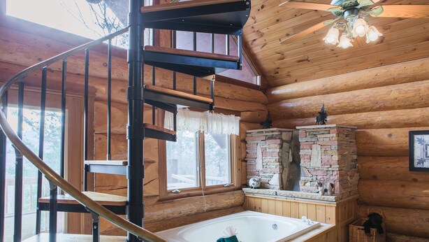 Interior de una cabaña de madera con escalera de caracol, un ventanal grande, chimenea de piedra y una bañera de madera.