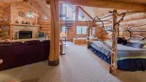 Interior de una cabaña de troncos con chimenea de piedra, una cama de madera con cabecera rústica, un sofá de cuero y una escalera en espiral.