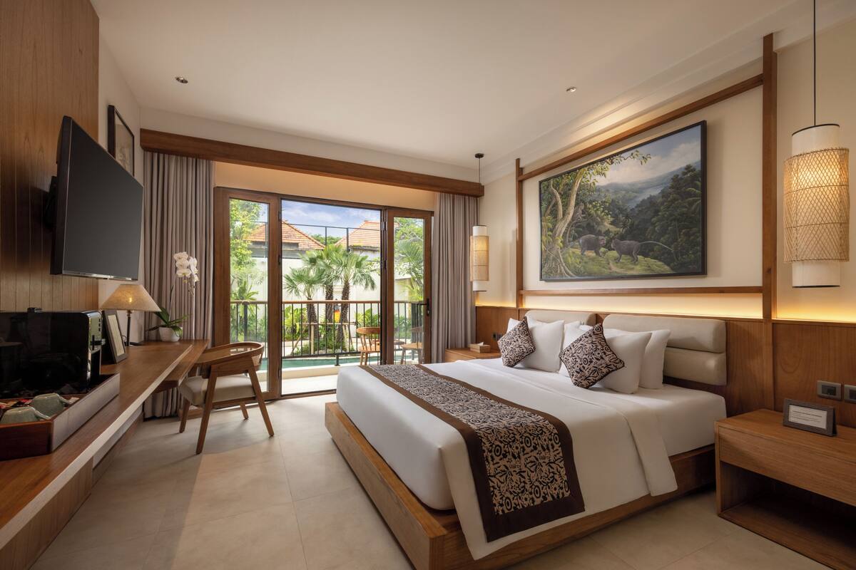 A hotel room with a large bed, a desk with a TV, a chair, a balcony with a view of palm trees, and a painting on the wall.