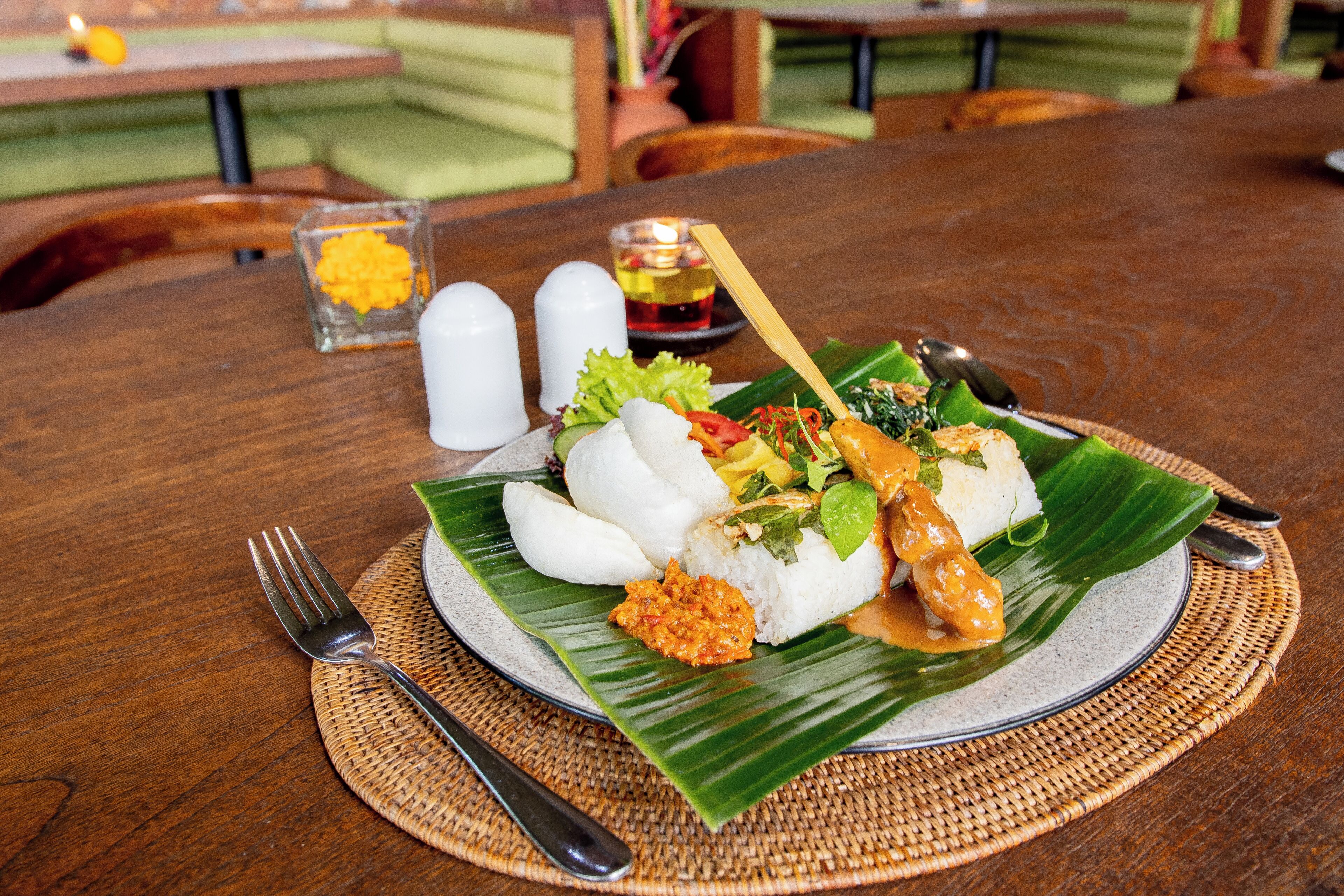 A meal served on a banana leaf with white rice, vegetables, and a sauce, on a wooden table with a fork and spoon.