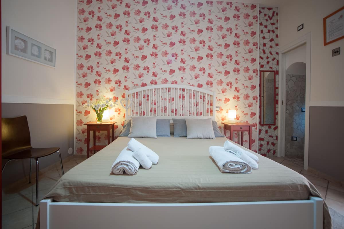 A neatly made bed with two rolled towels, a floral wallpaper, a white headboard, and a small table with a vase of flowers.