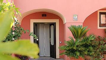 Un edificio de fachada rosa, una puerta con cortina y varias macetas con plantas en el suelo y en el balcón.