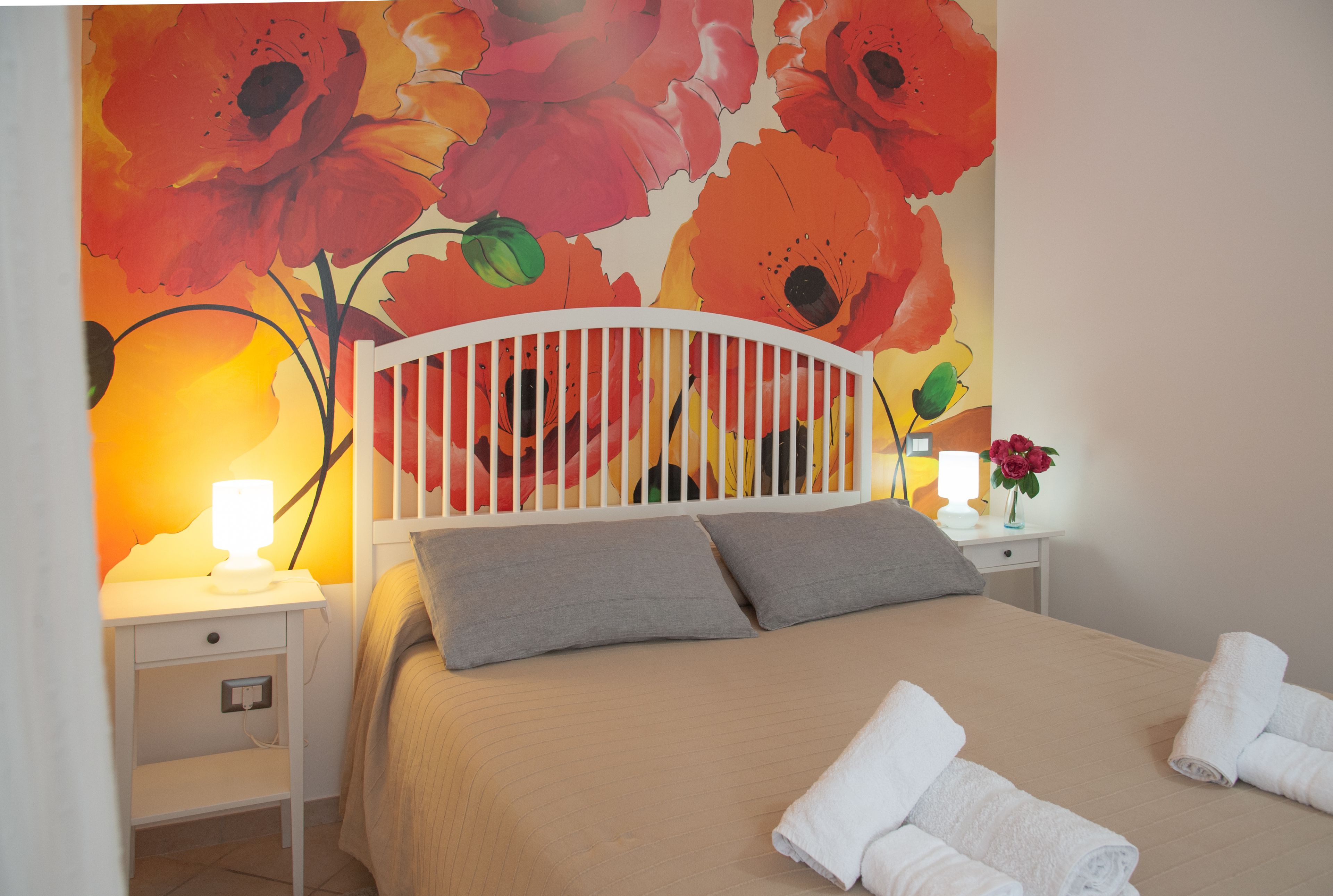 Una habitación de hotel con una cama, almohadas, una mesita de noche con lámpara y una pared con un mural floral.