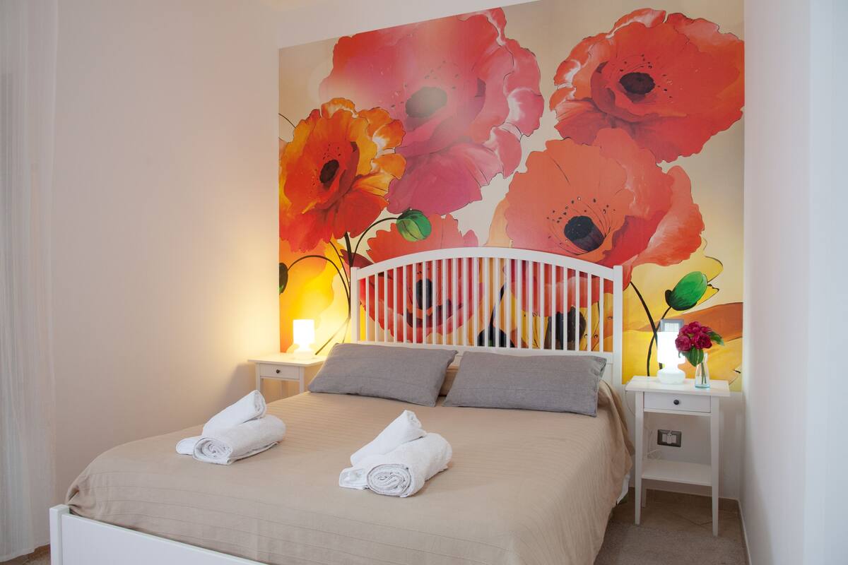 A neatly made bed with two rolled towels, a white headboard, and a wall with a large floral painting.