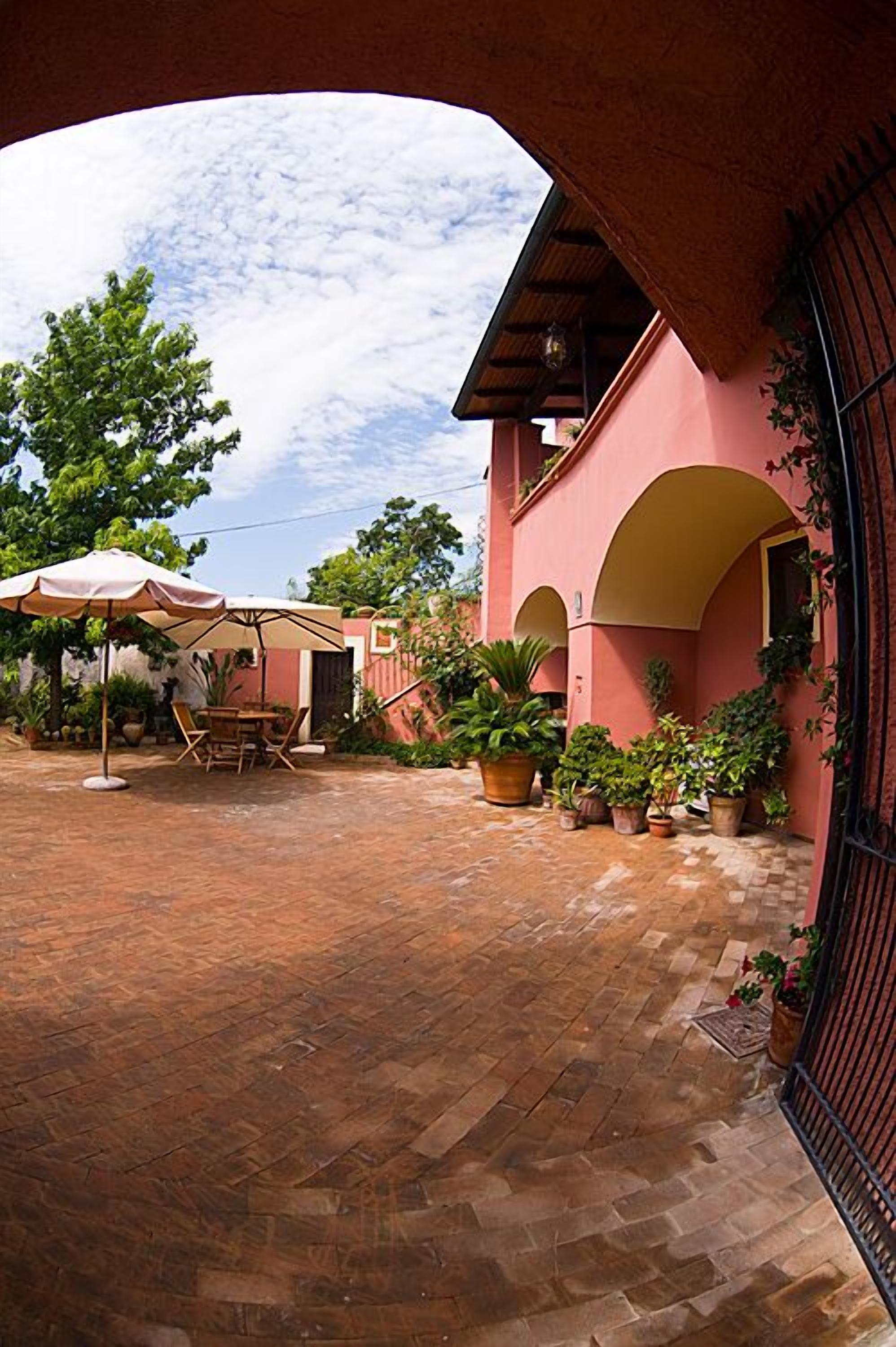A courtyard with a brick floor, a table with chairs, and an umbrella. There are potted plants and a building with arches.