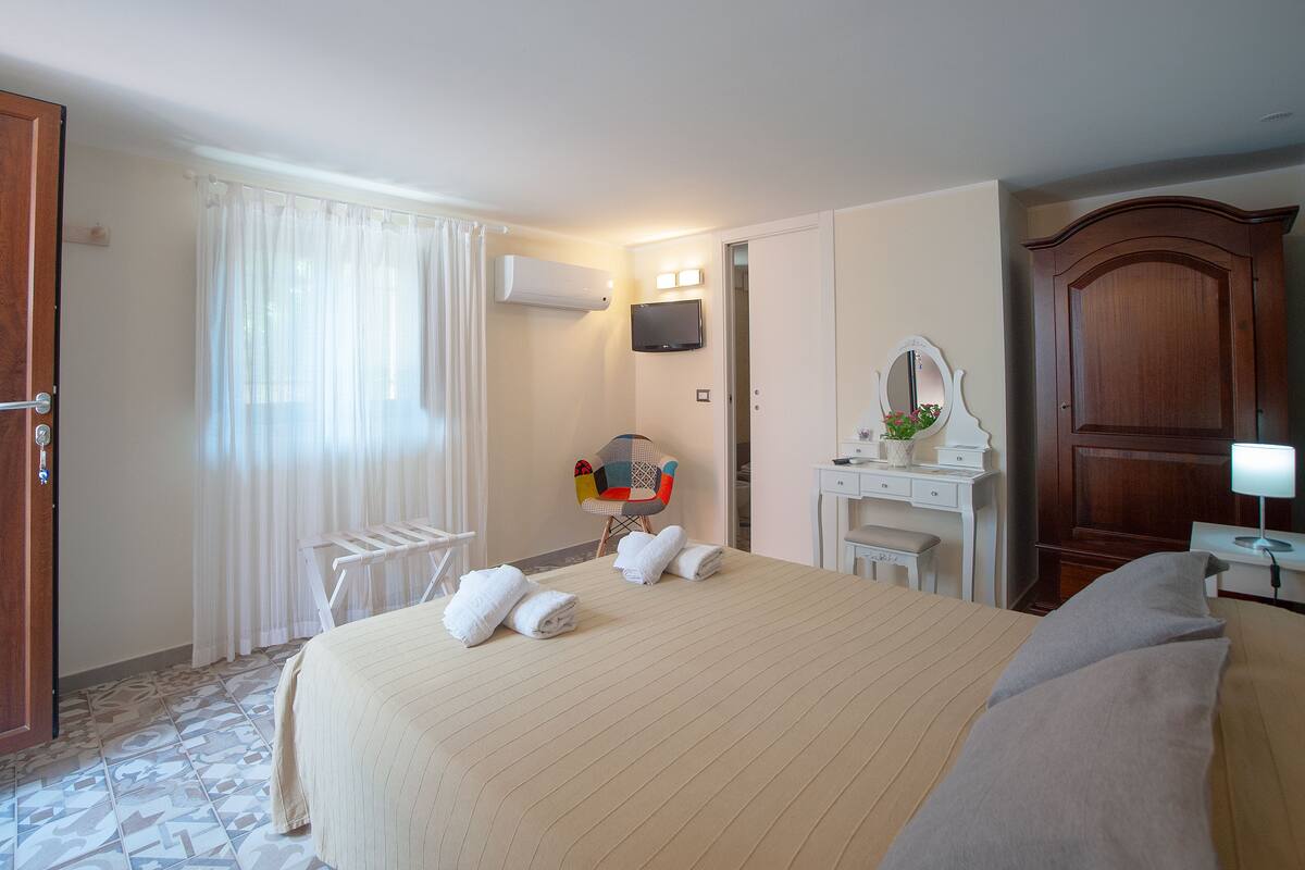 A hotel room with a large bed, a desk, a chair, a TV, and a wardrobe.