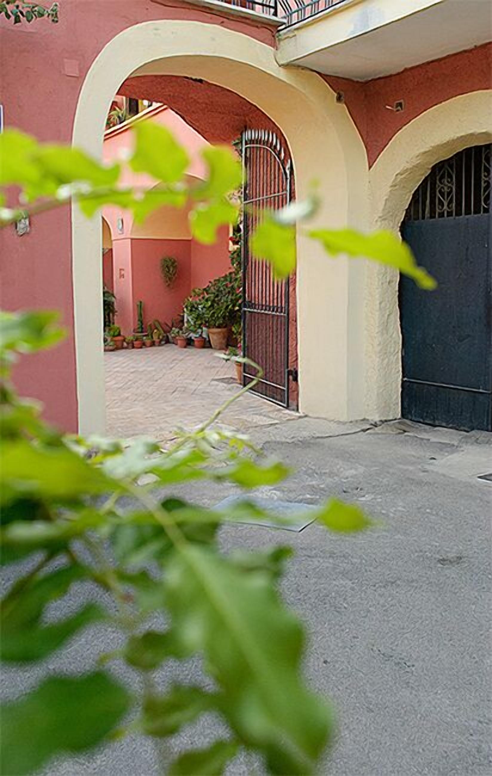 A courtyard with a gated entrance, potted plants, and a building with a pink facade and white arches.