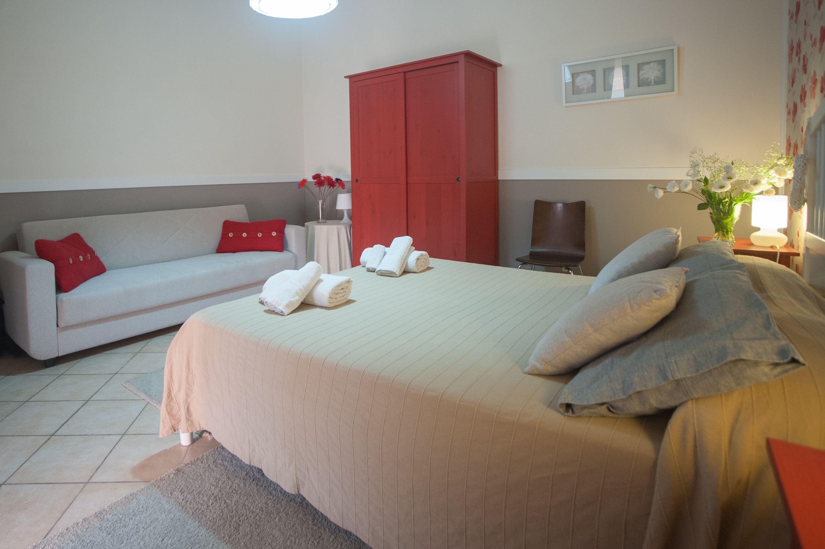 A hotel room with a bed, a sofa, a red wardrobe, a small table with a vase of flowers, and a lamp.