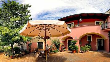 A courtyard with a table and chairs under an umbrella, surrounded by potted plants and a building with arches.