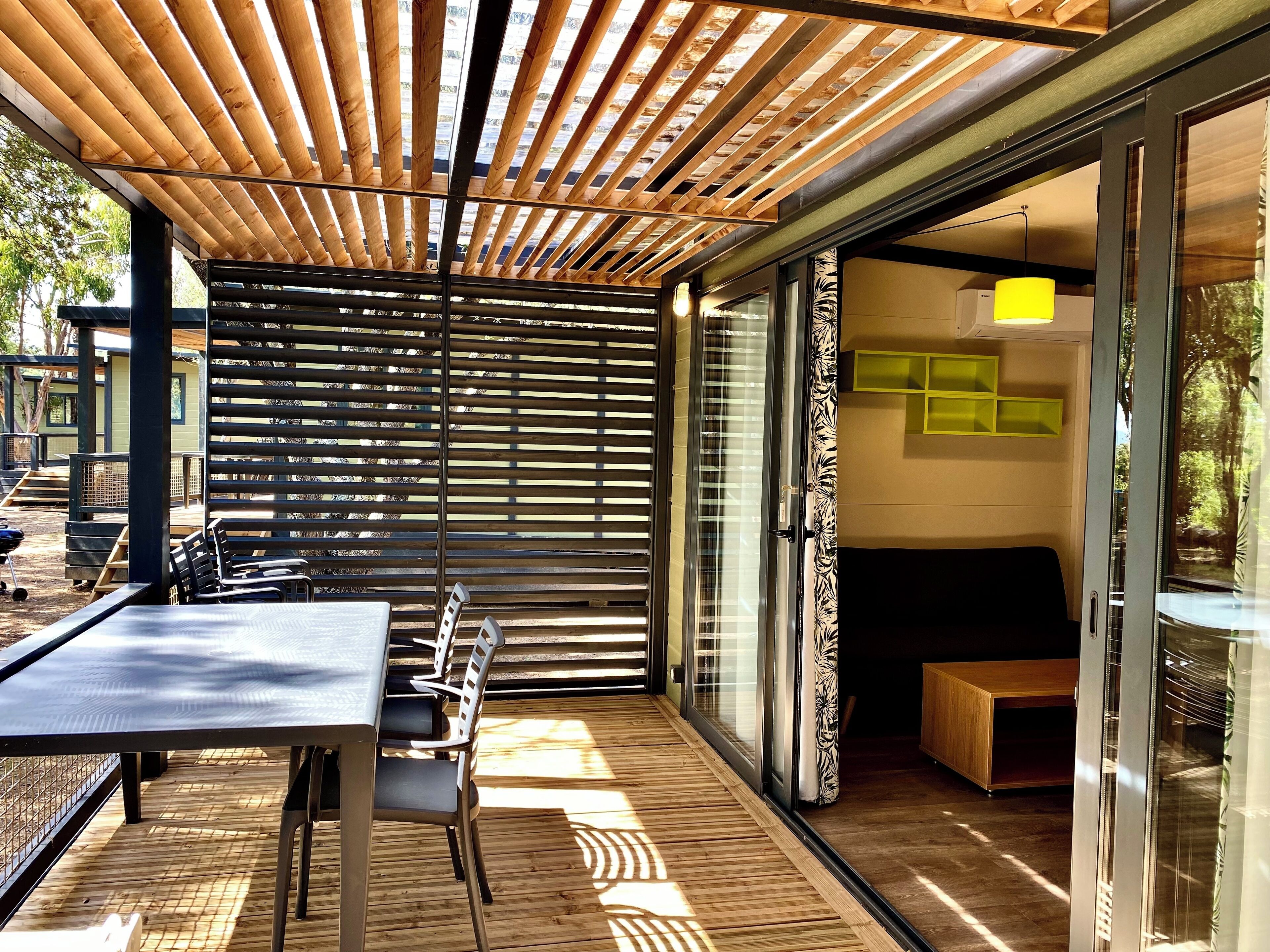 An outdoor patio with a wooden pergola, a dining table with chairs, and a glass door leading to an interior space with a sofa and a yellow lamp.