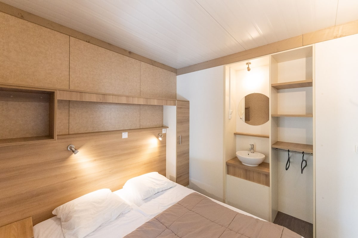 Une chambre moderne comprenant un lit, des rangements intégrés en bois, une table de chevet et une salle de bain avec un lavabo et un miroir.