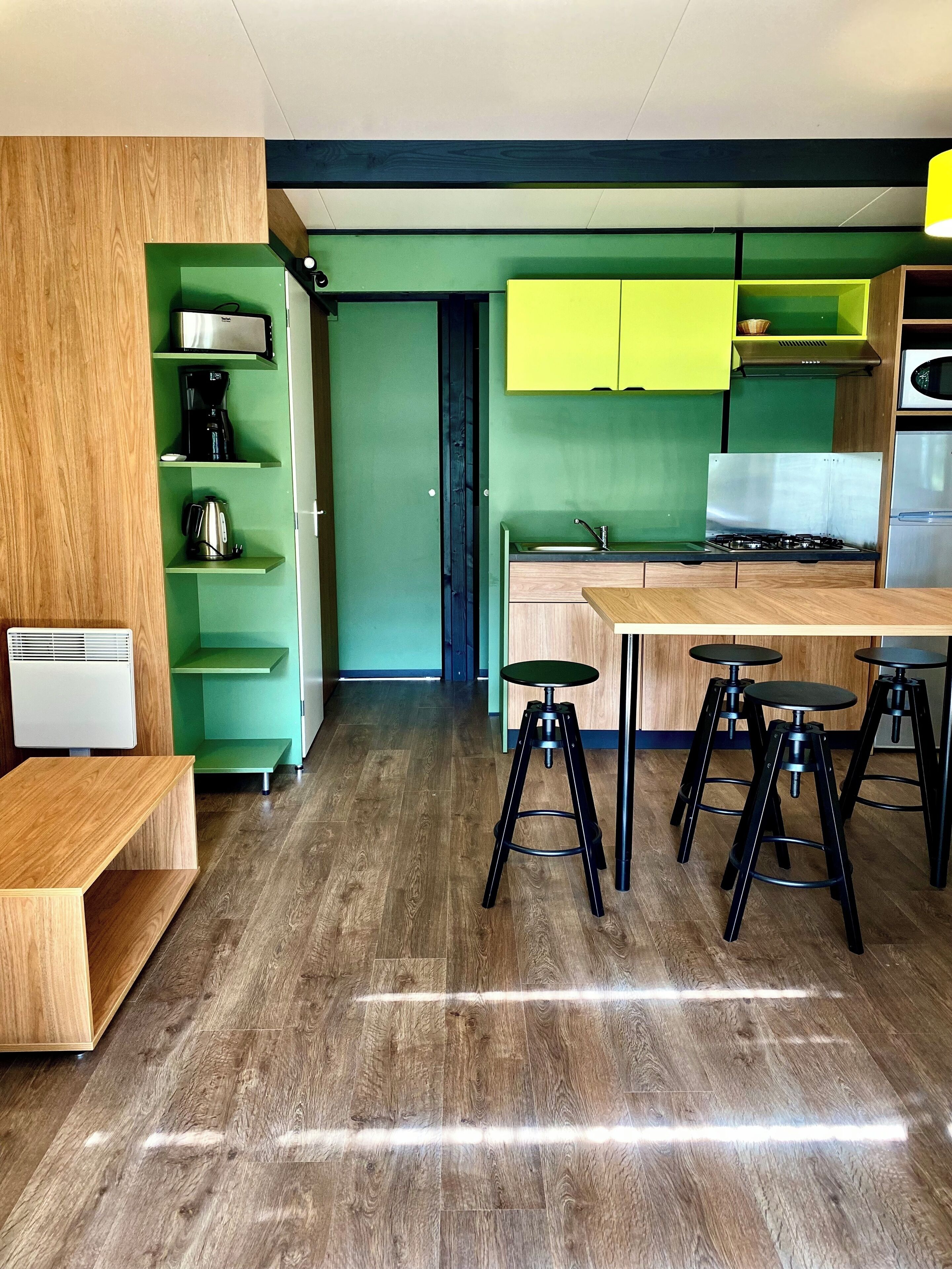 Une cuisine moderne avec des meubles de couleur verte, un plan de travail en bois et une cuisinière à gaz. On y trouve trois tabourets et un banc.