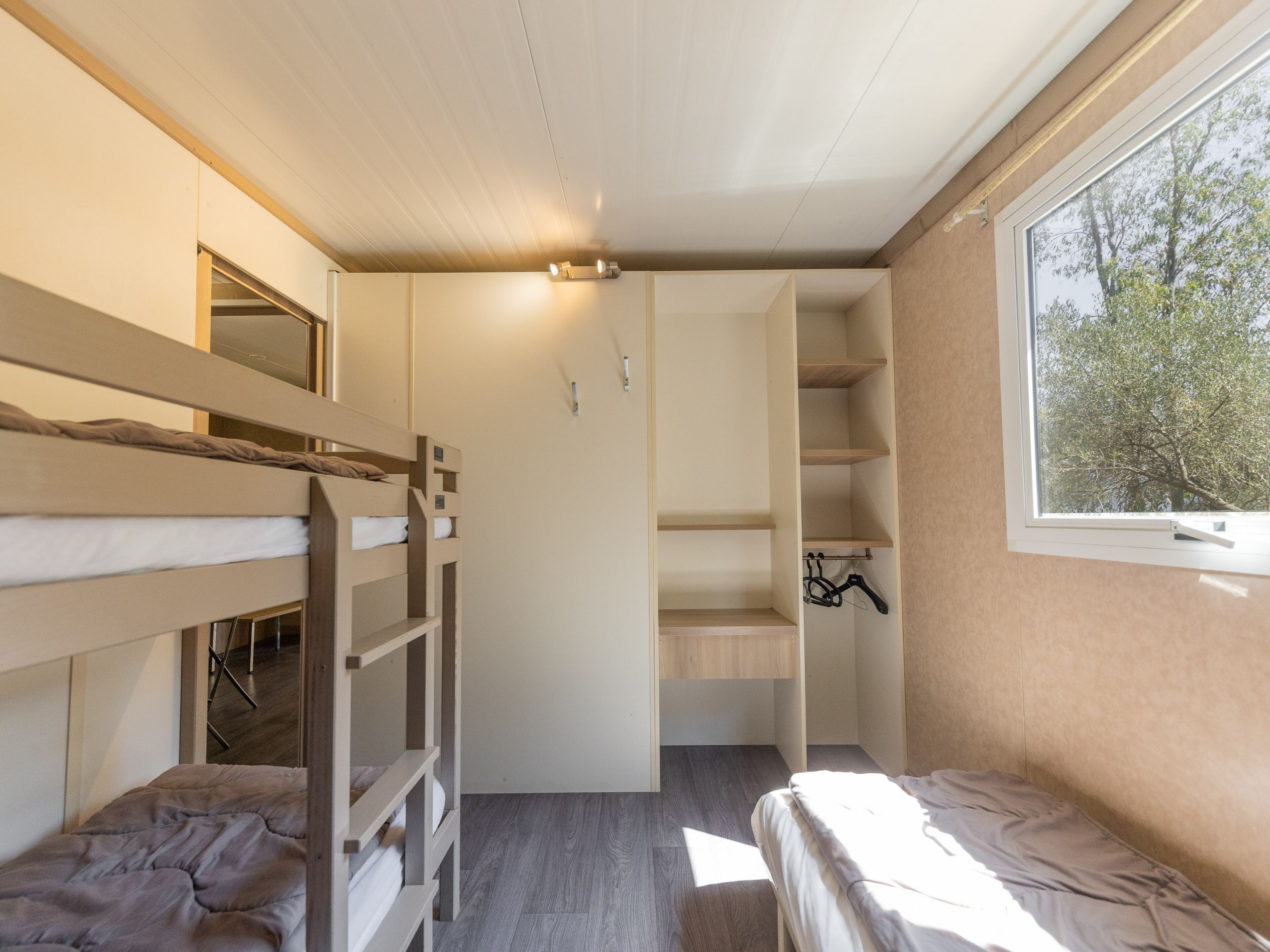 A dormitory room with bunk beds, a wardrobe, and a window with a view of trees.
