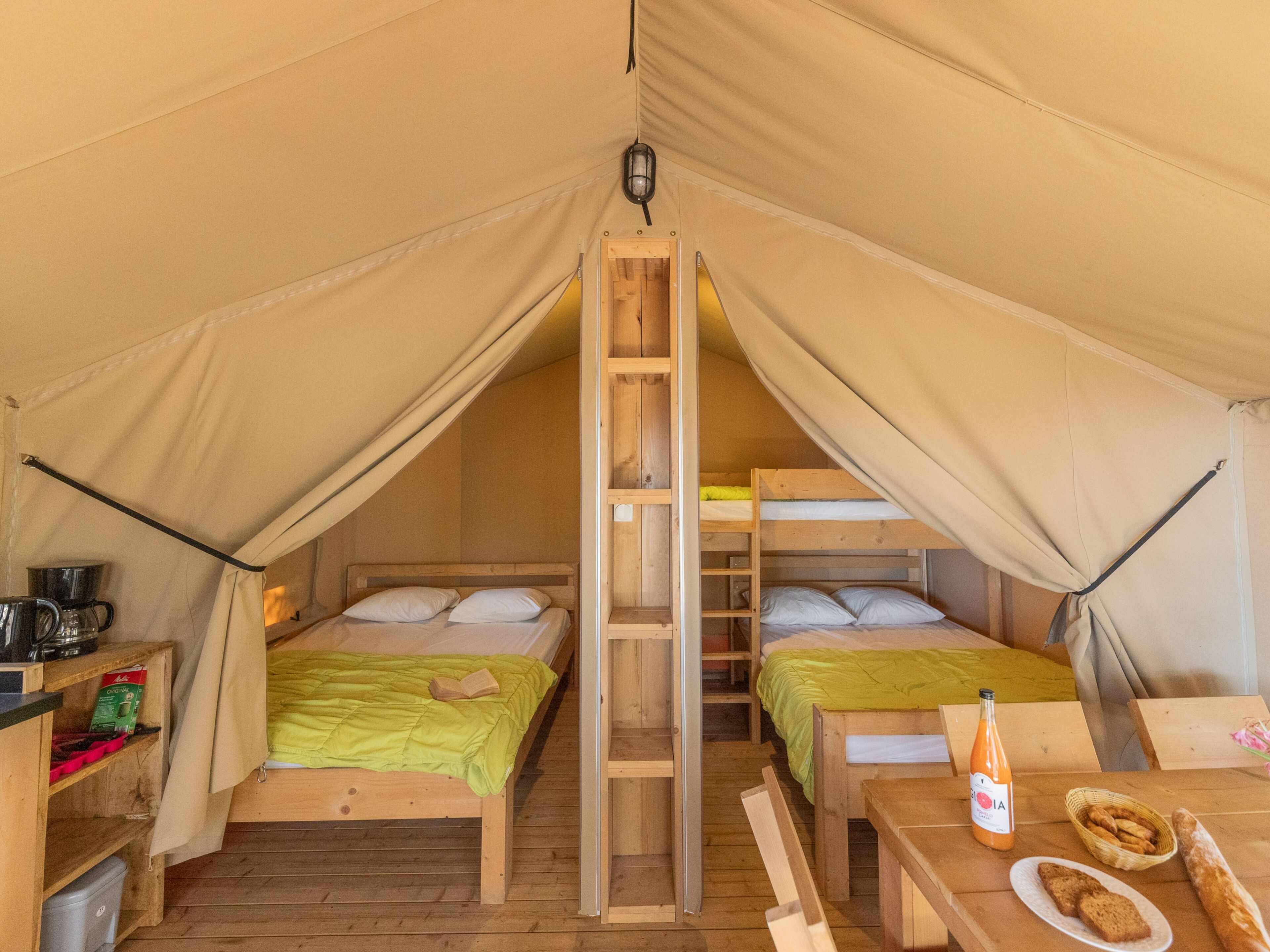 A tent with two bunk beds, a ladder, a table with bread and a bottle, and a shelf with a kettle.