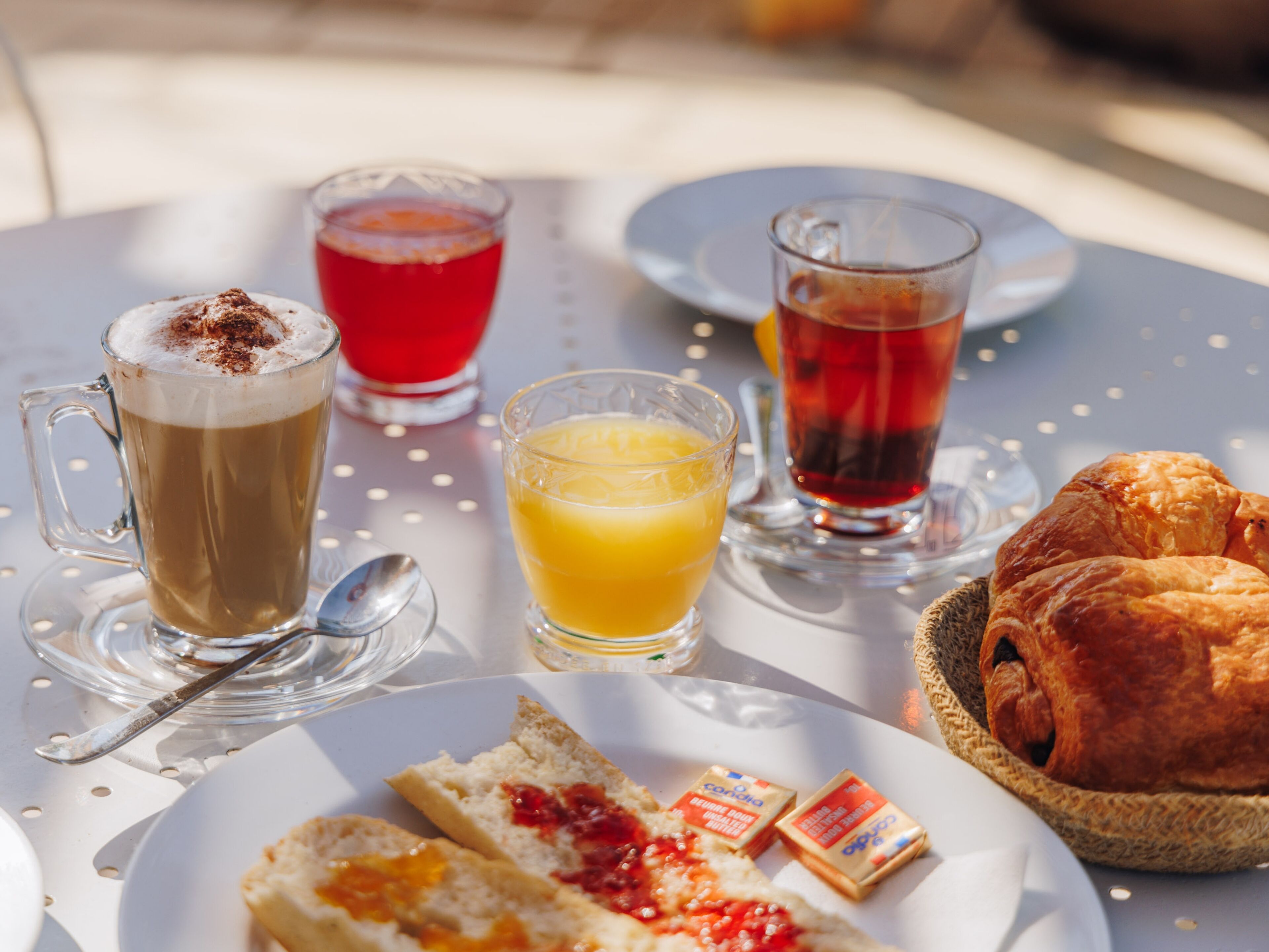 Une table avec une tasse de café, un verre de jus d’orange, une tasse de thé, des croissants et des tartines de confiture.
