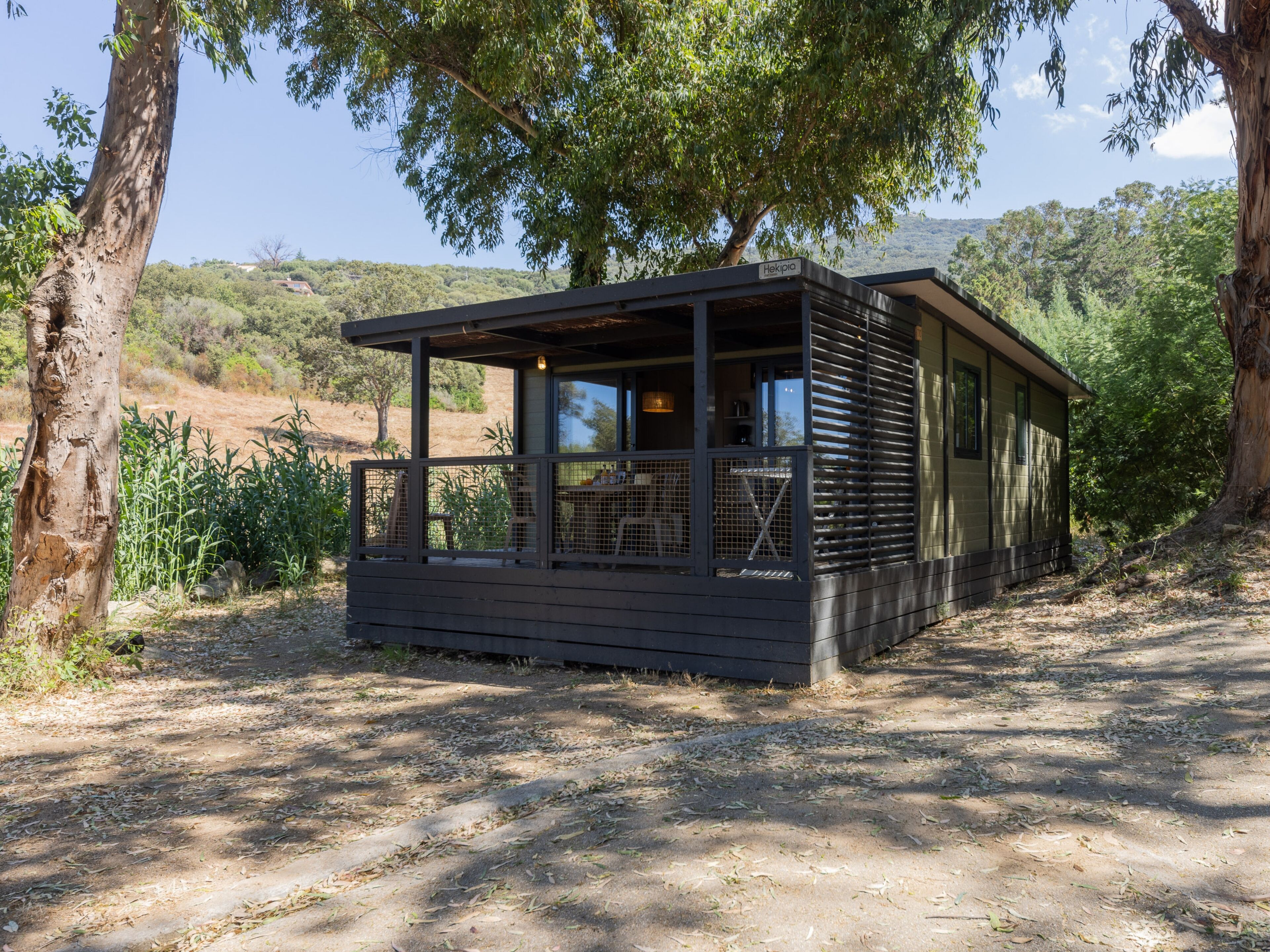 A small cabin with a covered porch and a dining area, surrounded by trees and a hilly landscape.
