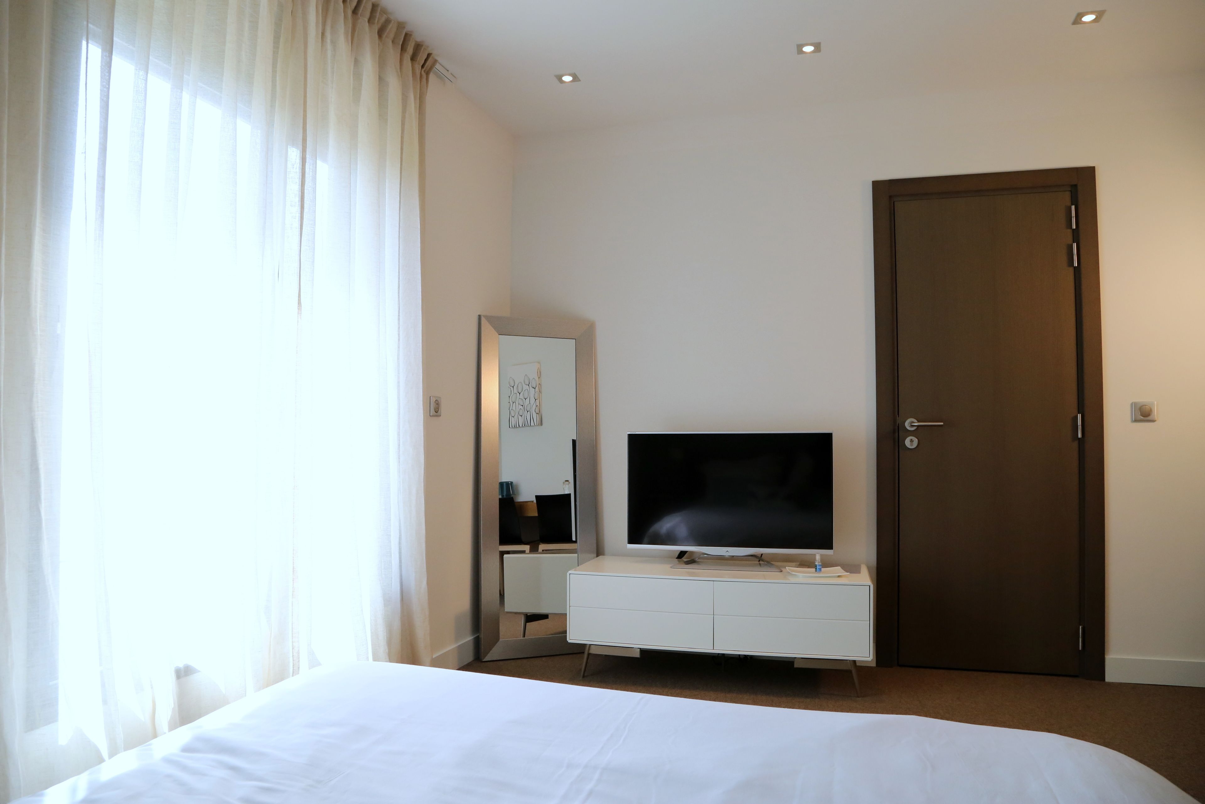 A hotel room with a bed, a TV on a white dresser, a door, and a window with curtains.