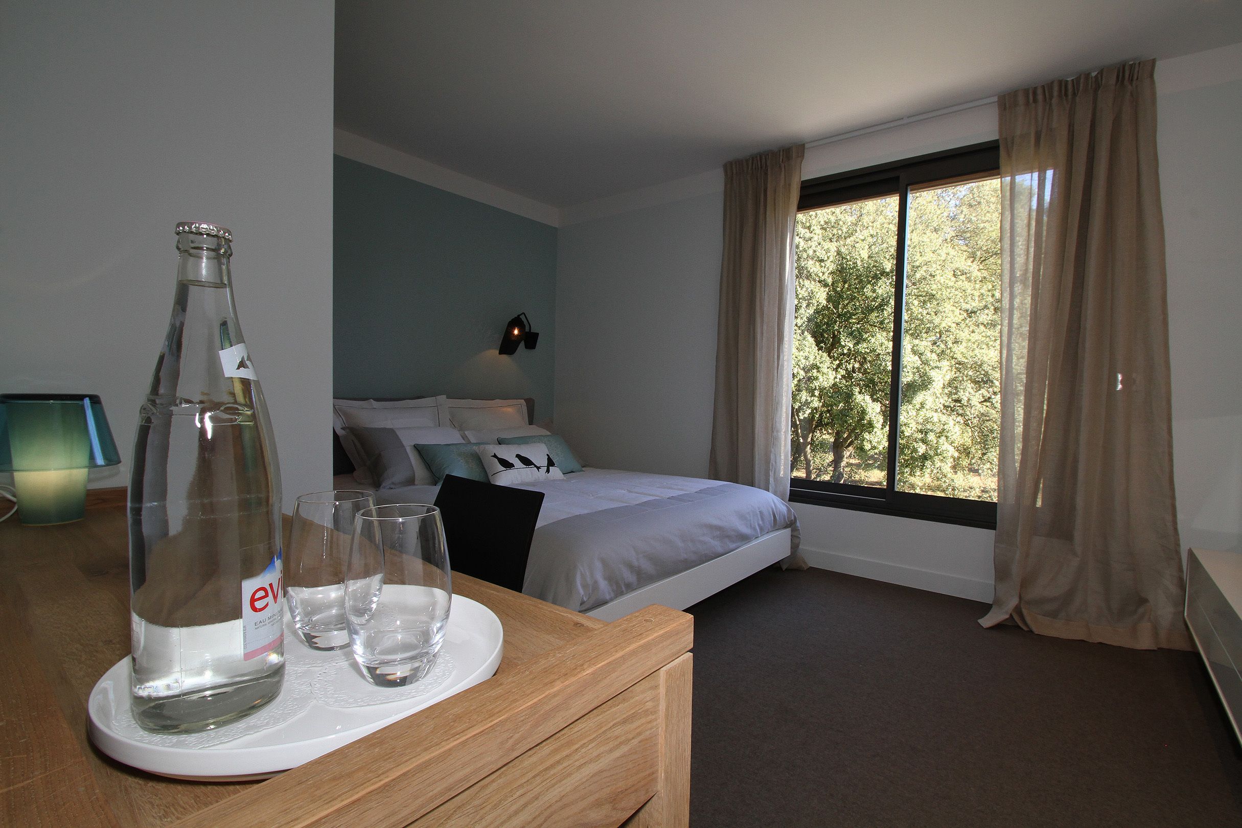 A hotel room with a bed, a bedside table with a bottle and glasses, a lamp, and a window with curtains.