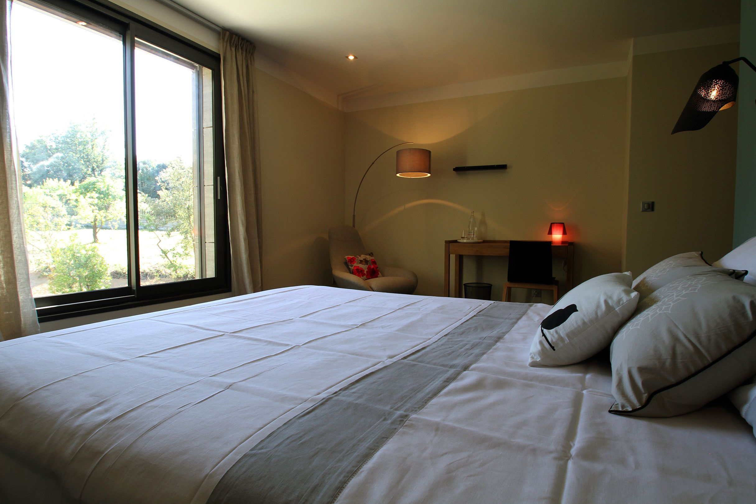 A neatly made bed with white linens, a chair with a red pillow, a bedside table with a lamp, and a window with curtains.