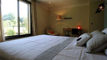 A neatly made bed with white linens, a chair with a red pillow, a bedside table with a lamp, and a window with curtains.