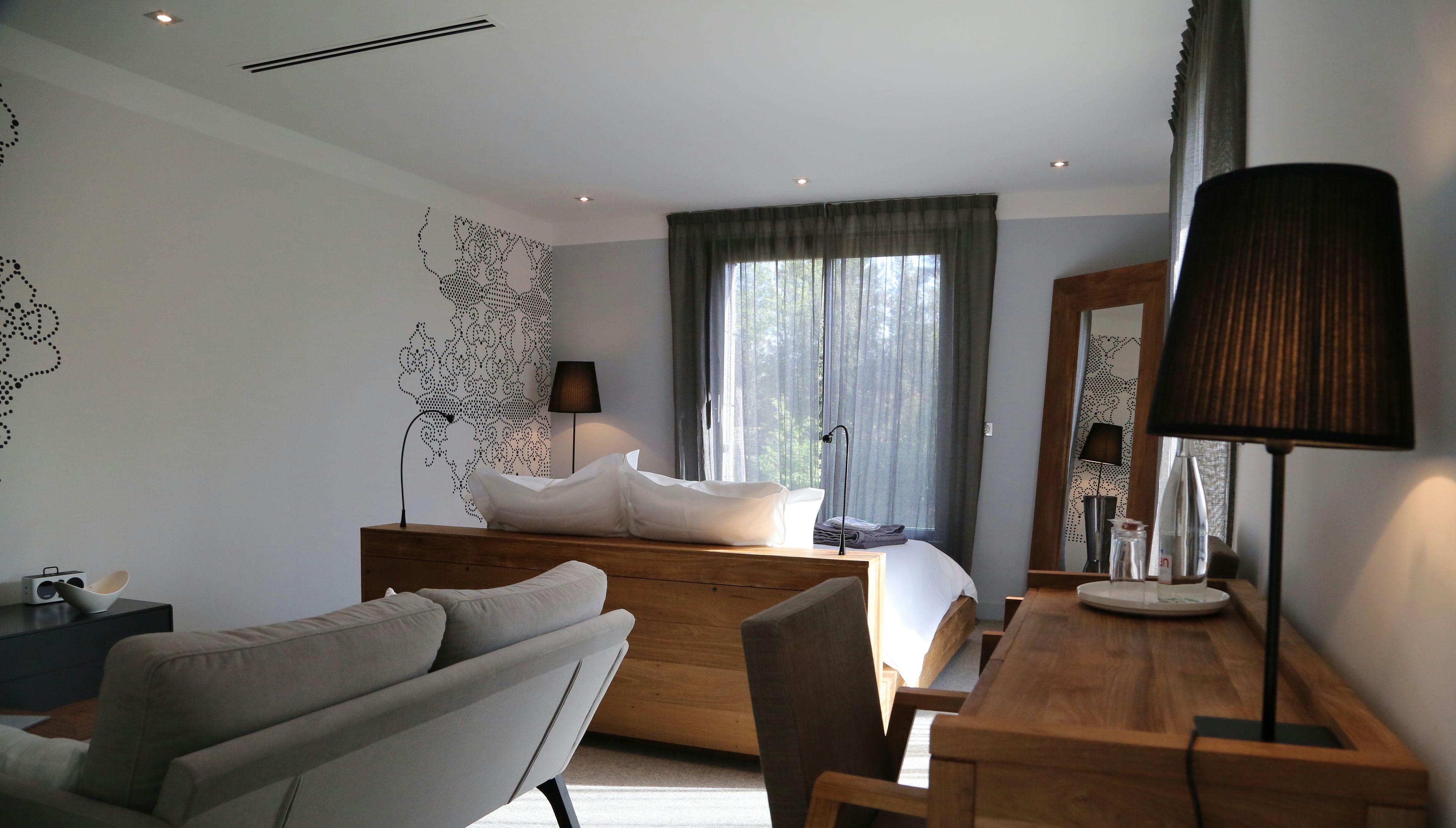 A modern bedroom with a wooden bed, a grey sofa, a wooden desk, and a large window with curtains.