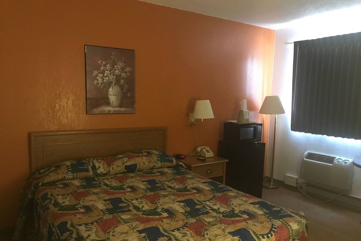 A hotel room with a bed, a desk with a telephone and a lamp, a television, and an air conditioning unit.