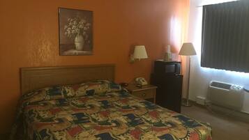 A hotel room with a bed, a desk with a telephone and a lamp, a television, and an air conditioning unit.