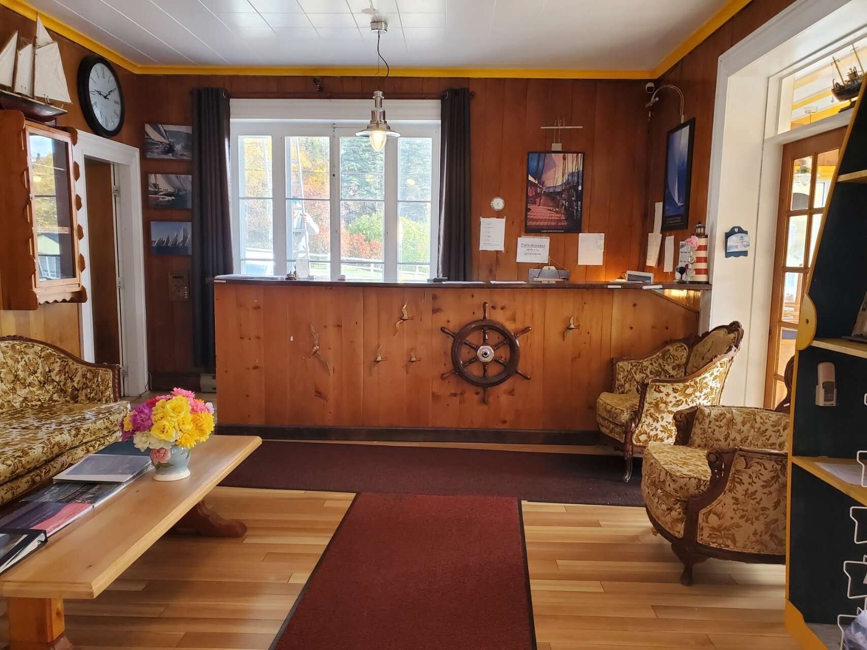 Una camera accogliente con pareti in legno, una scrivania di legno, due divani decorati e un ornamento a forma di timone da nave, con una grande finestra che si affaccia su alberi.