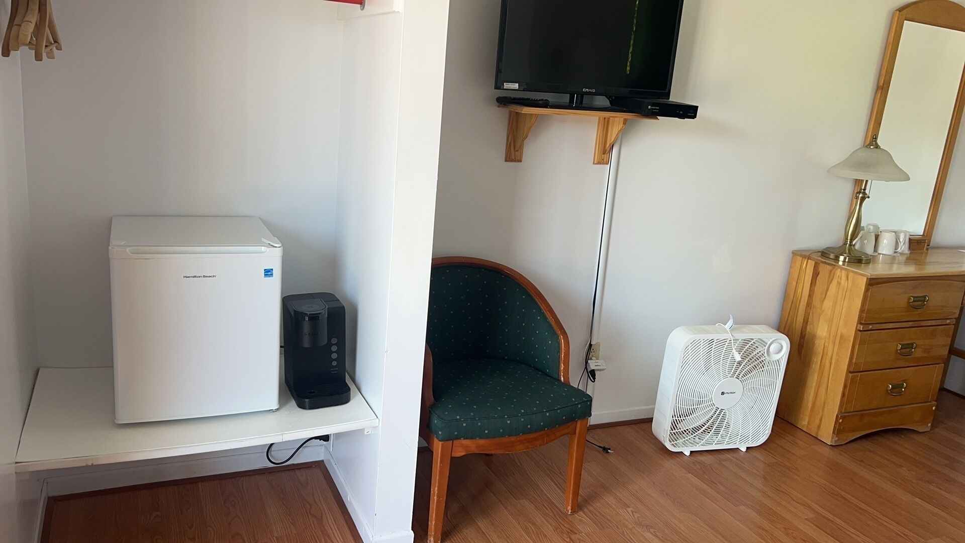 Une chambre d’hôtel avec un réfrigérateur blanc, une petite machine à café, un fauteuil vert, un ventilateur blanc, une commode en bois et un téléviseur fixé au mur.