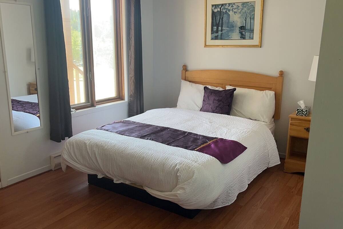A neatly made bed with a white and purple bedspread, a wooden headboard, and a nightstand with a tissue box.