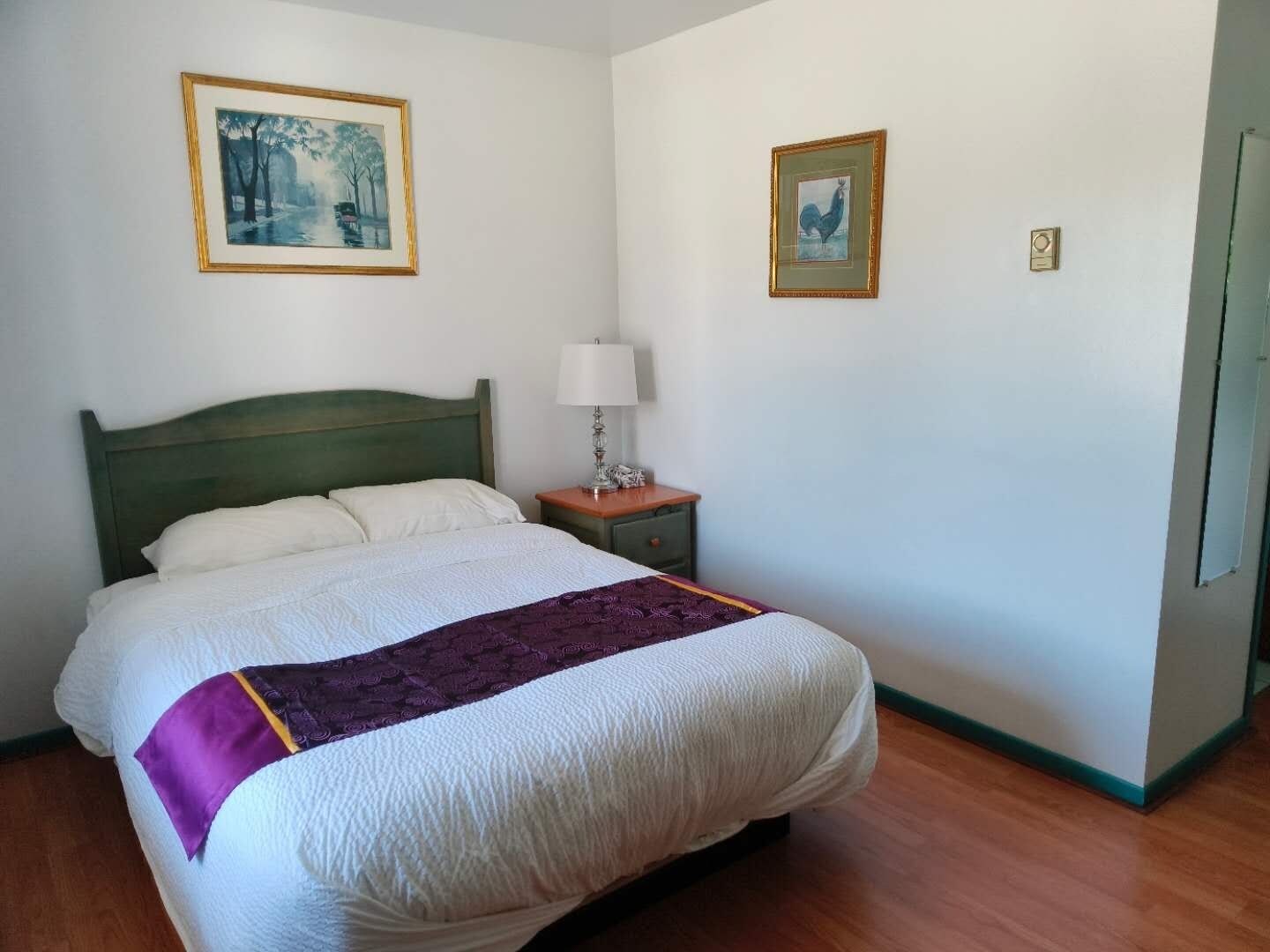 Uma cama bem arrumada com cabeceira verde, mesa de cabeceira com abajur e duas fotografias emolduradas na parede.