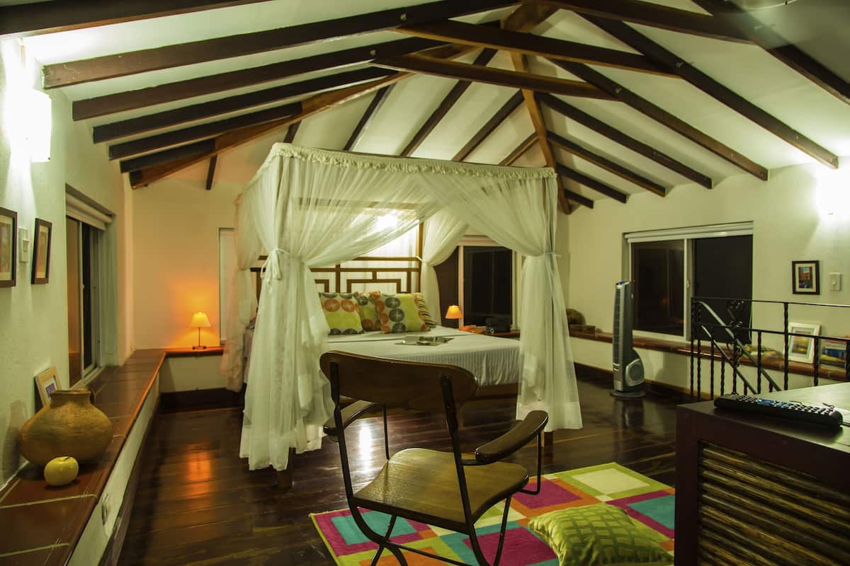 Un dormitorio con cama con dosel, una silla de madera, una alfombra colorida y un ventanal grande.