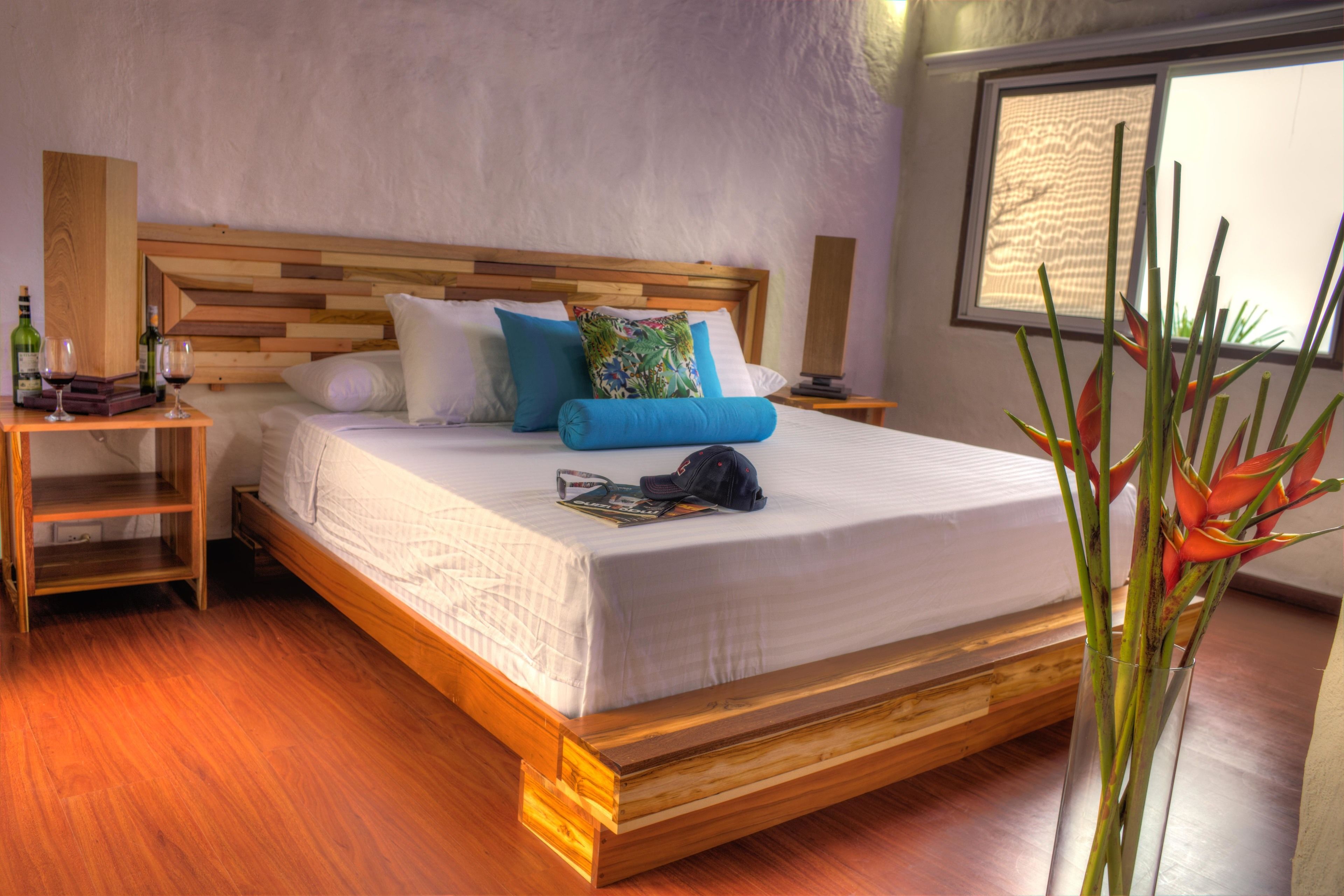 Una cama bien hecha con cabecera de madera, una mesita de noche con una botella y vasos, y una ventana con vistas al exterior.