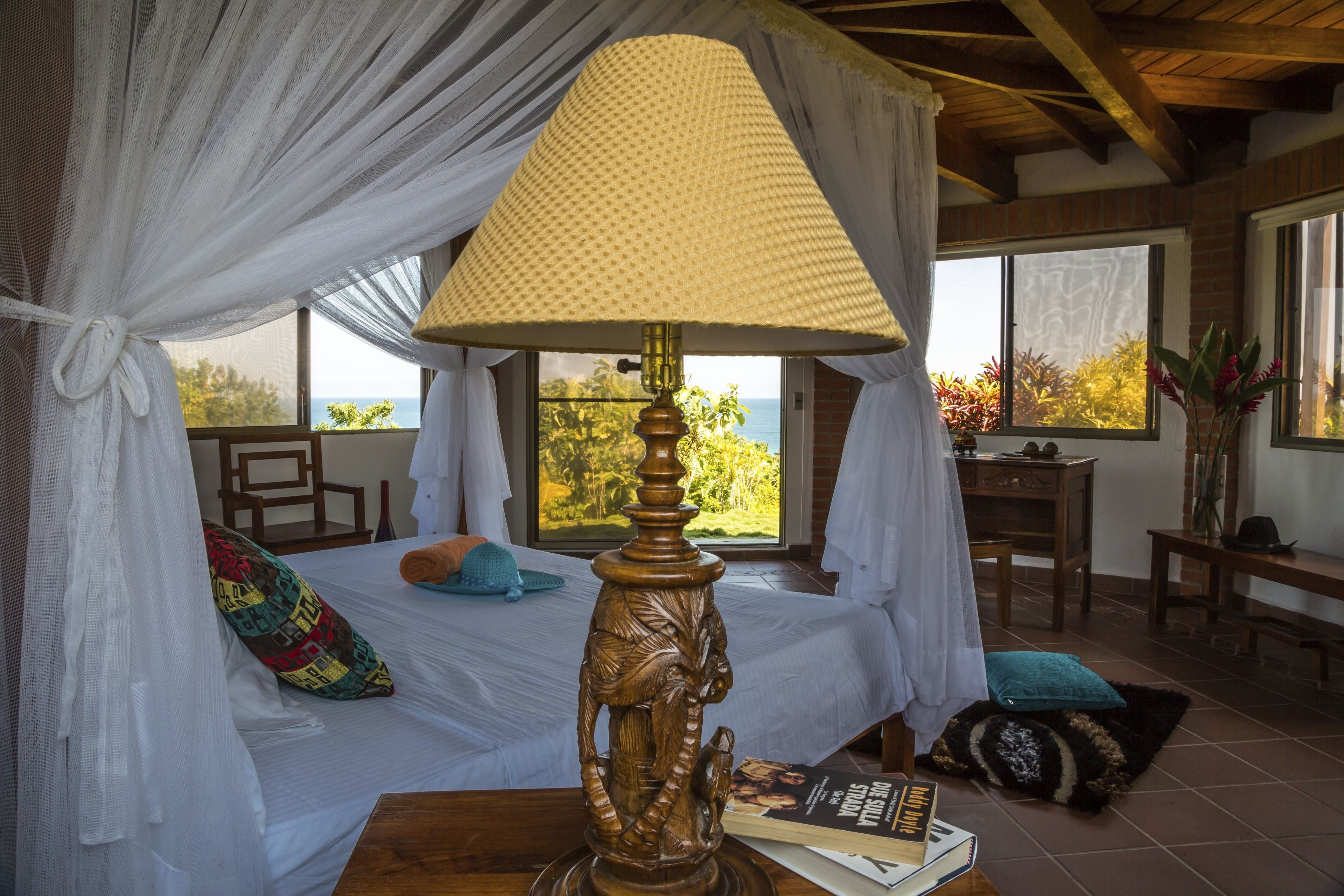 A bedroom with a bed under a canopy, a wooden lamp, a desk, and a view of the outdoors.