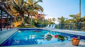 Relax by the serene pool surrounded by lush greenery and palm trees, with lounge chairs and a refreshing fruit bowl nearby.