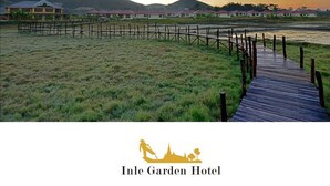 A wooden boardwalk leading to a row of buildings with red roofs, surrounded by grassy fields and hills in the background.