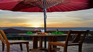 An outdoor dining table with chairs, a red umbrella, and a view of the sunset over a body of water.